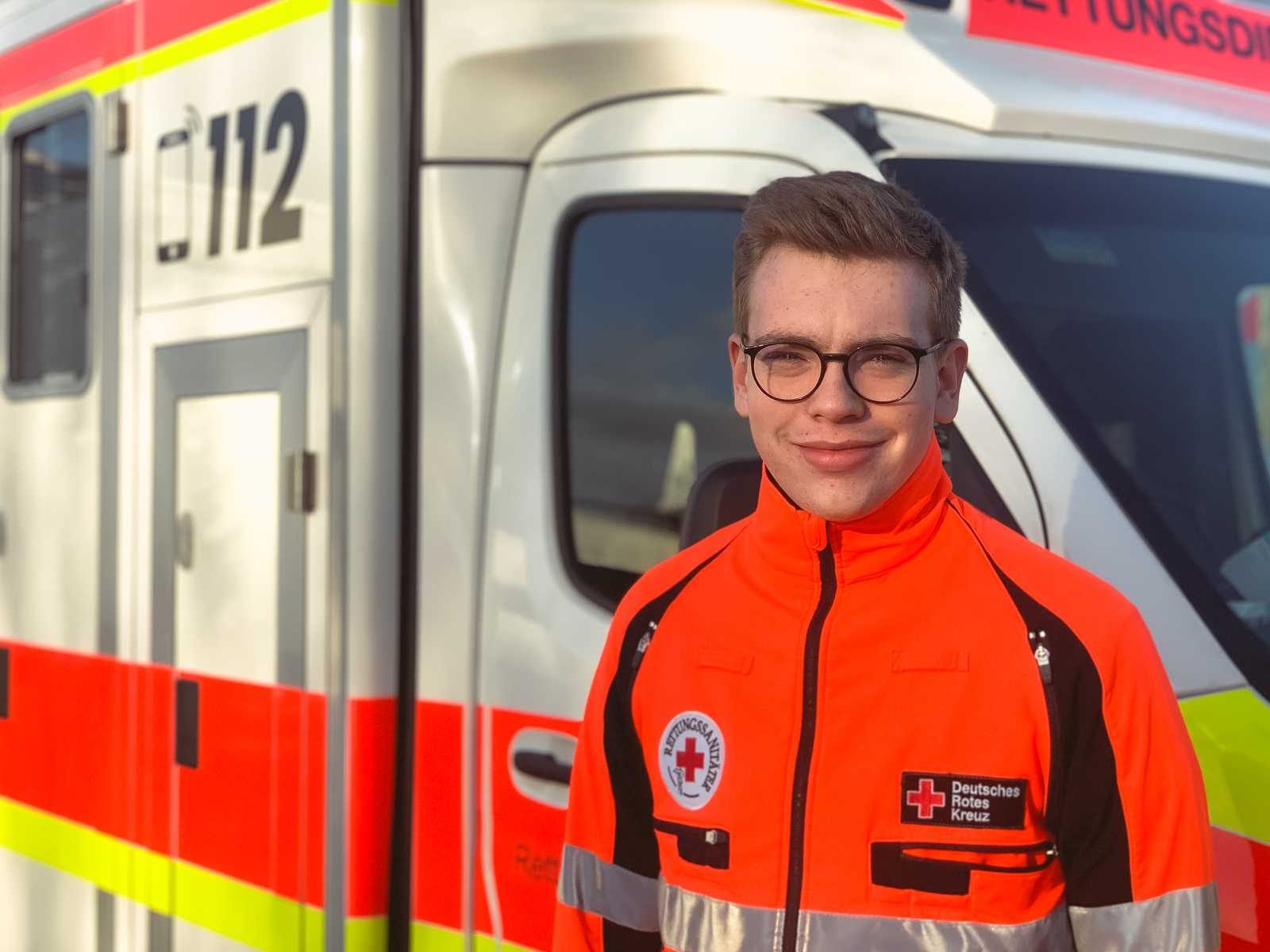 Ein junger Rettungssanitäter mit Brille steht in seiner Arbeitsjacke vor einem Rettungswagen.