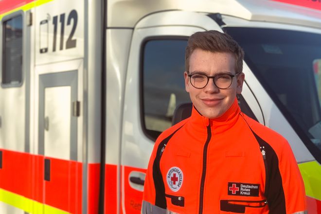 Ein junger Rettungssanitäter mit Brille steht in seiner Arbeitsjacke vor einem Rettungswagen.