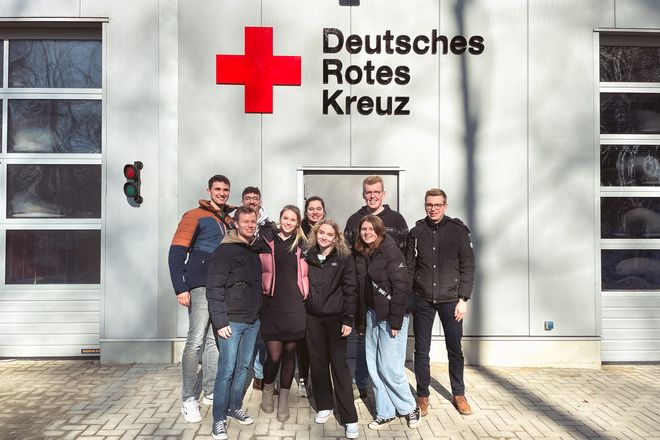 Eine Gruppe von neun jungen Rettungssanitäter:innen steht zwischen zwei Fahrzeugtoren einer Rettungswache. Die Sonne scheint und im Hintergrund sieht man das Logo des Deutschen Roten Kreuzes.